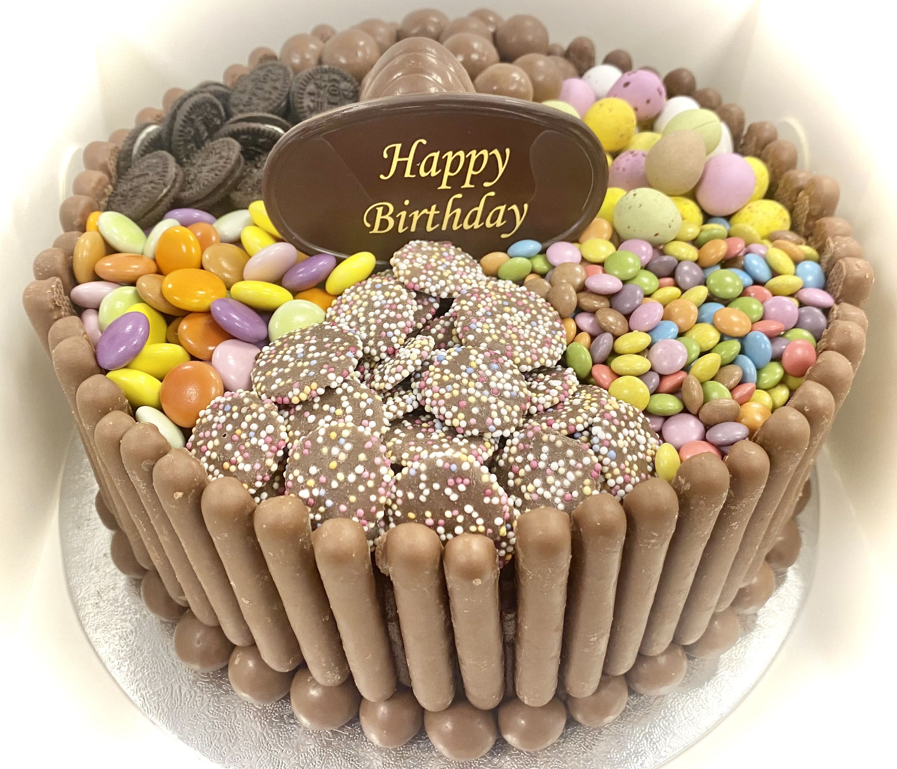A decorated birthday chocolate cake is a chocoholic's dream with an assortment of candies and chocolates. The cake is surrounded by chocolate sticks and topped with treats, including sprinkle-covered chocolates, colorful candy-coated chocolates, mini chocolate eggs, and Oreo cookies. A chocolate plaque inscribed with 'Happy Birthday' is placed centrally on top.